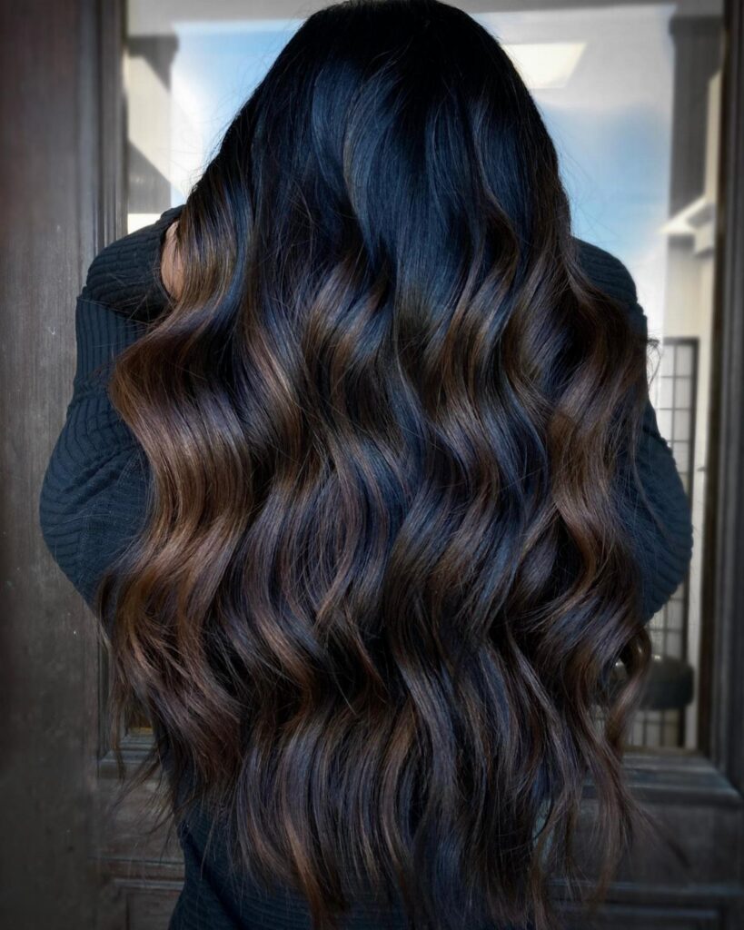 Long black wavy hair with subtle brown balayage highlights over a dark sweater