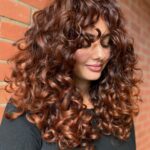 Woman with long dark curls showing copper balayage against a brick wall