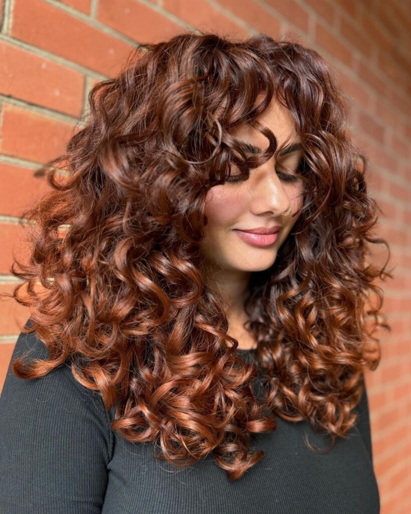 Woman with long dark curls showing copper balayage against a brick wall
